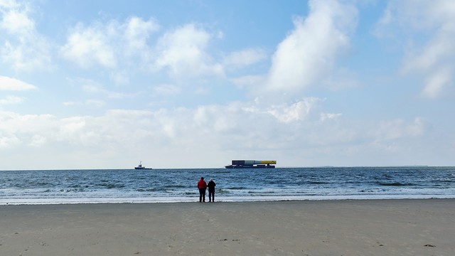 zwei Personen stehen an der Wasserkante und betrachten ein kleines Boot, das eine Plattform zieht, auf der ein sehr großes Rohr liegt. 