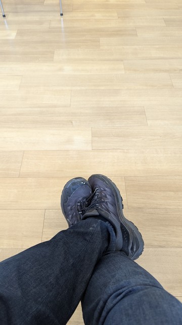 Fußboden in Parkettoptik, zwei gekreuzte Beine mit Jeans und schwarzen Turnschuhen.