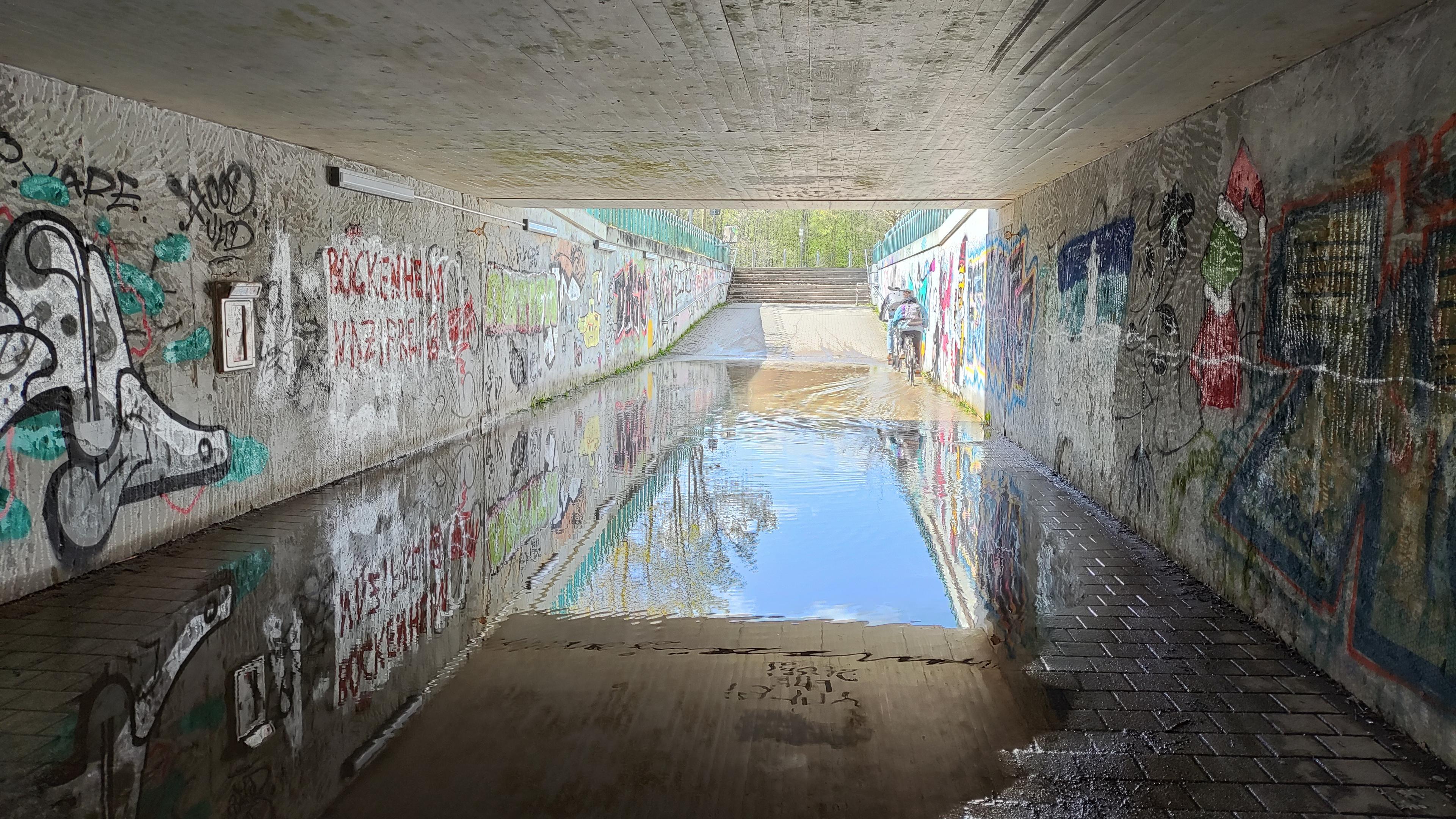 unterführung mit besprühten Wänden auf deren Boden das Wasser auf ganzer breite steht in dem sich das Tageslicht spiegelt