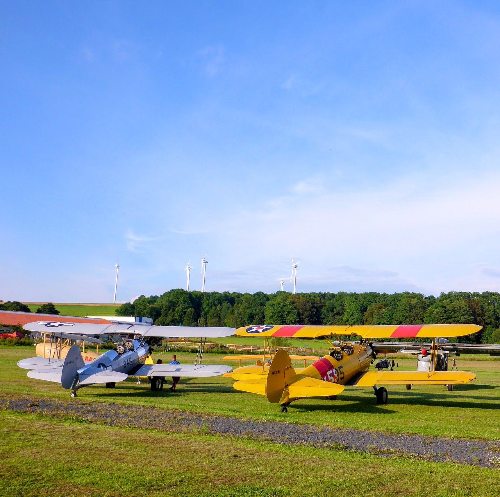 zwei historische Flugzeuge auf einer großen Wiese und dahinter etwas Wald und ein strahlend blauer Himmel. 