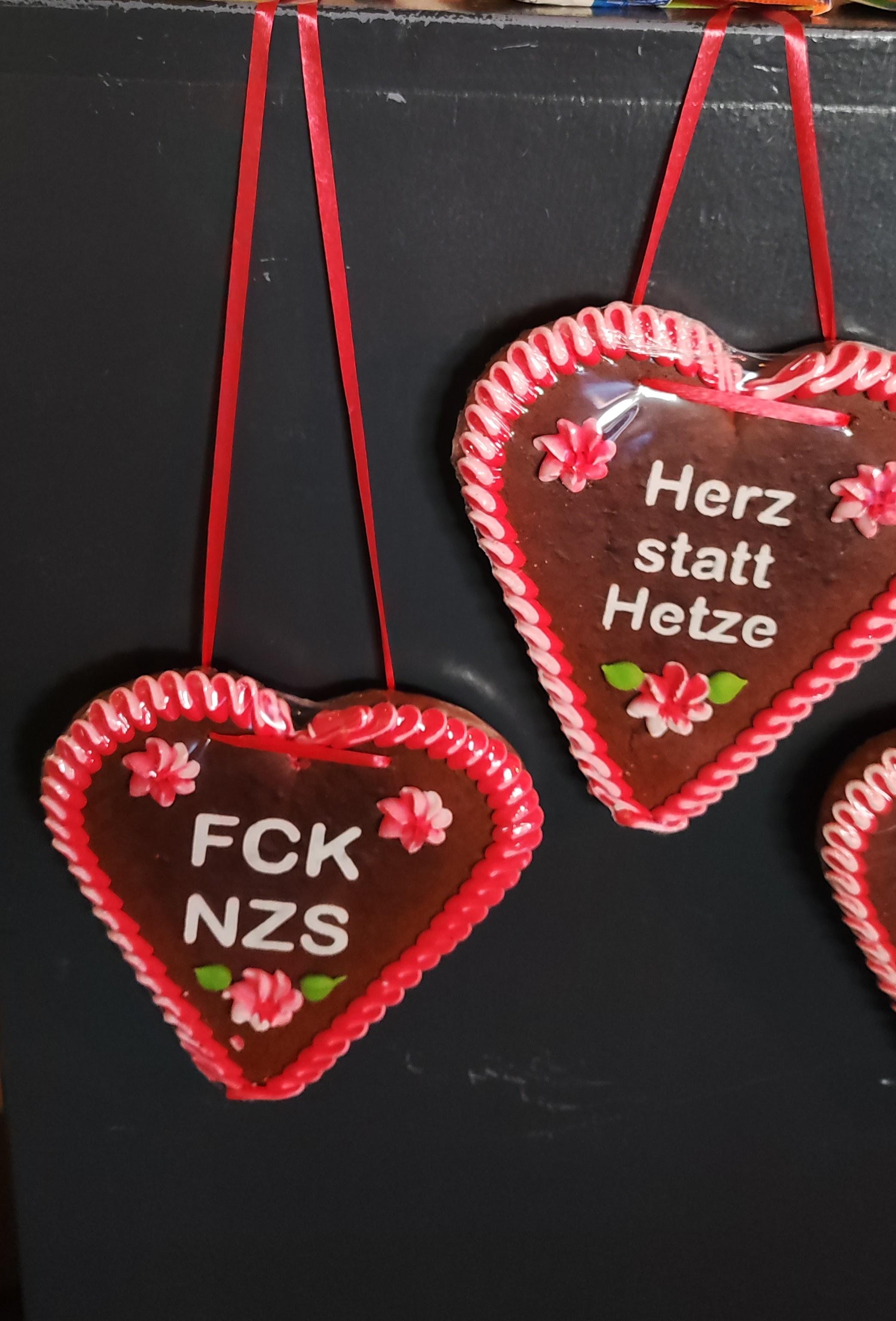 Zwei herzförmige Lebkuchen mit rotem Rand und kleinen rosa Zuckerdekorationen hängen an roten Bändern vor einem dunklen Hintergrund. Auf dem linken Lebkuchen steht in weißen Großbuchstaben 'FCK NZS'. Auf dem rechten Lebkuchen steht in weißen Buchstaben 'Herz statt Hetze'.