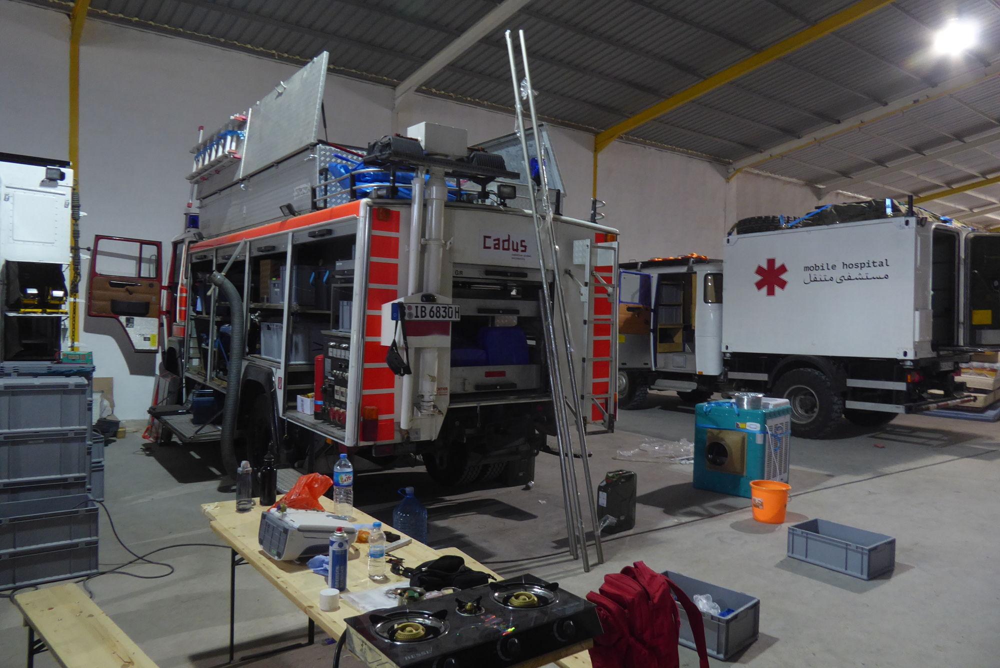 garage mit einem großen Krankenwagen drin der die Geräteräume offen hat. Viel gerät werkzeug kocher usw liegt überall rum