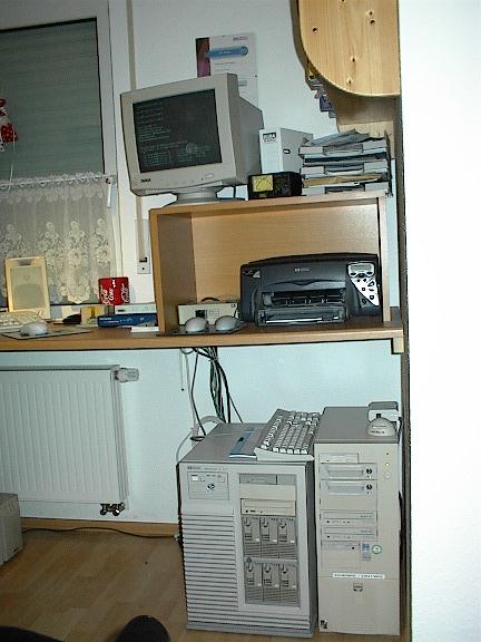 An early-2000s home computer setup. On the floor, an HP NetServer LH Pro server beside a self-built Pentium PC. Above them sits an HP PhotoSmart P1000 printer with a 15-inch Targa CRT monitor displaying a text-based console. A beige keyboard, mouse, speakers, stacks of CDs and manuals complete the workspace. The photo captures a typical enthusiast's home lab from the SCSI and Windows NT era.